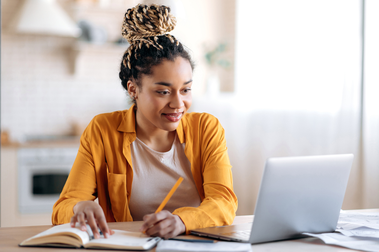 Focused cute stylish african american female student with afro dreadlocks, studying remotely from home in Social Media with Blazebridge Academy, using a laptop, taking notes on notepad during online lesson, e-learning concept, smiling