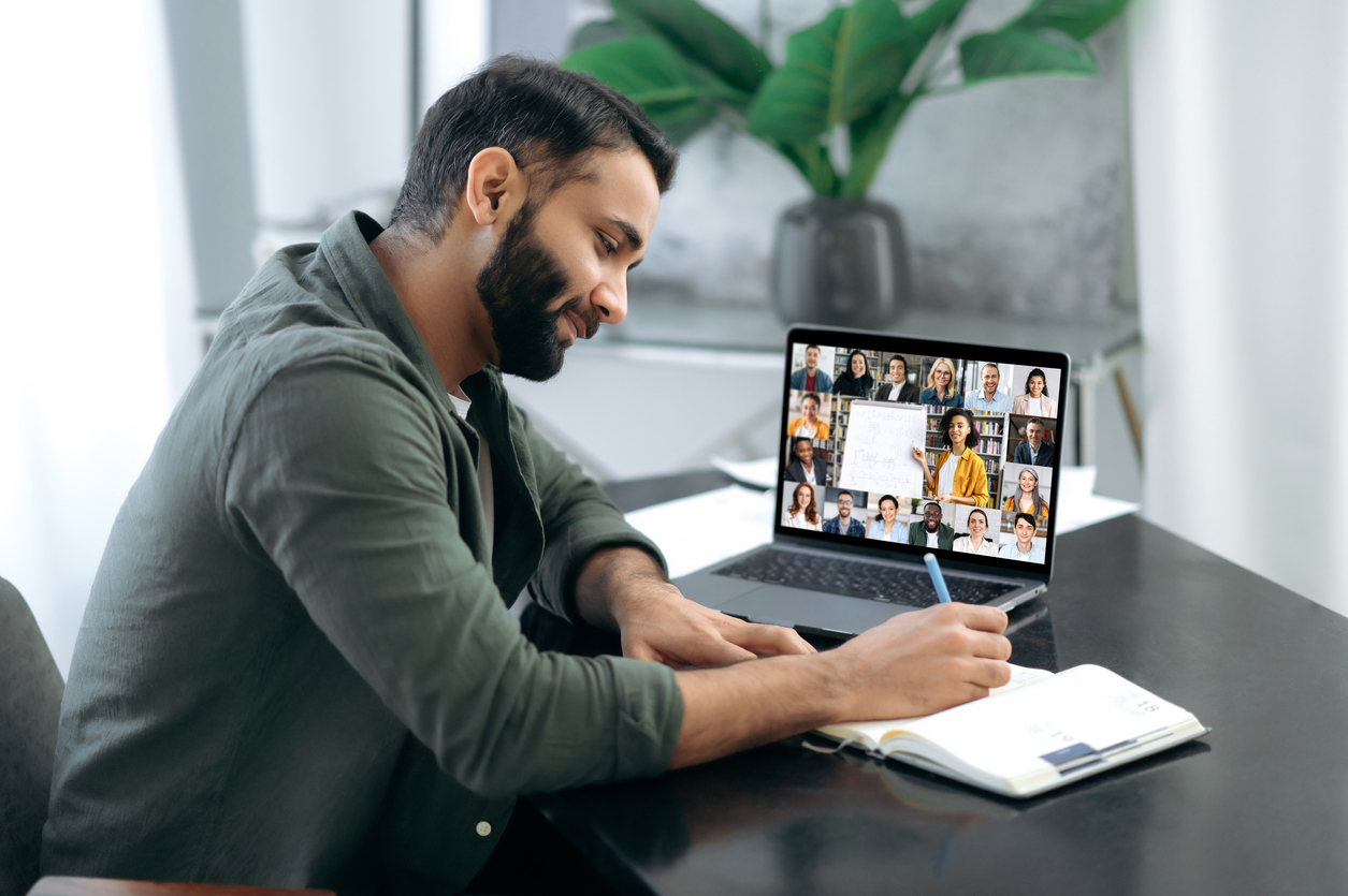 Side view of a successful smart guy listening to an online lecture in Social Media with Blazebridge Academy, taking notes in a notebook, on a laptop screen, a teacher and a group of multiracial people. Online training, webinar